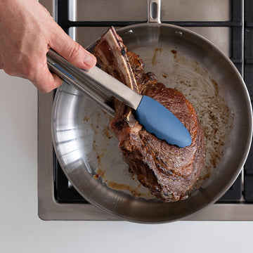 A blue pair of Misen Silicone Tongs holding and turning a well-seared ribeye steak over a stainless steel pan on a stovetop.